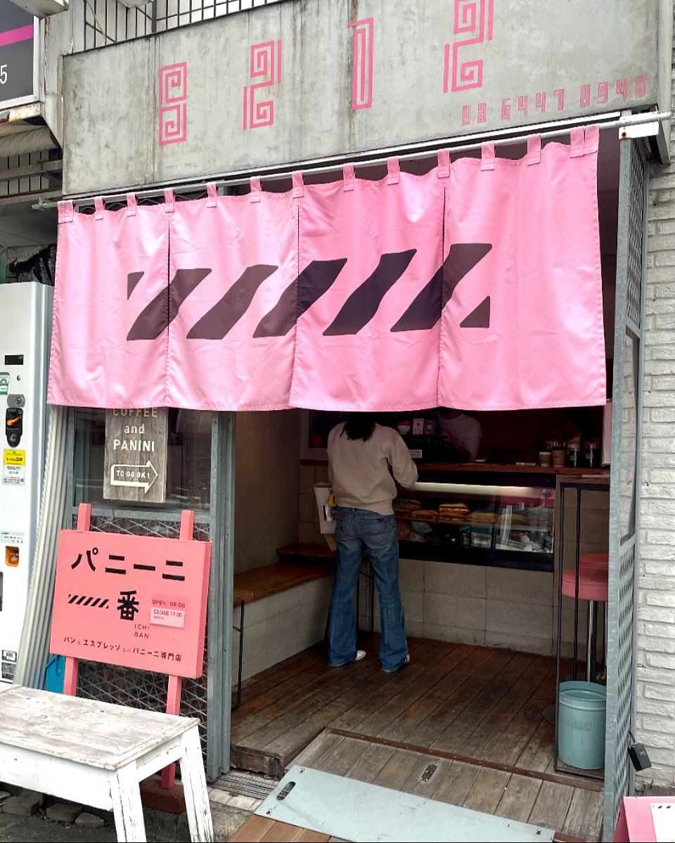 日本の東京都渋谷区千駄ケ谷にあるパニーニ一番の外観写真。
年季の入ったコンクリートに蛍光ピンクで文字が書かれている。のれん・看板の色も蛍光ピンク。